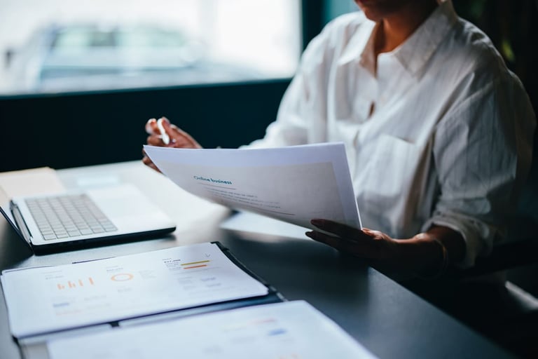 Pessoa analisando relatórios financeiros em ambiente de trabalho com computador e documentos, foco em negócios e planejamento estratégico.