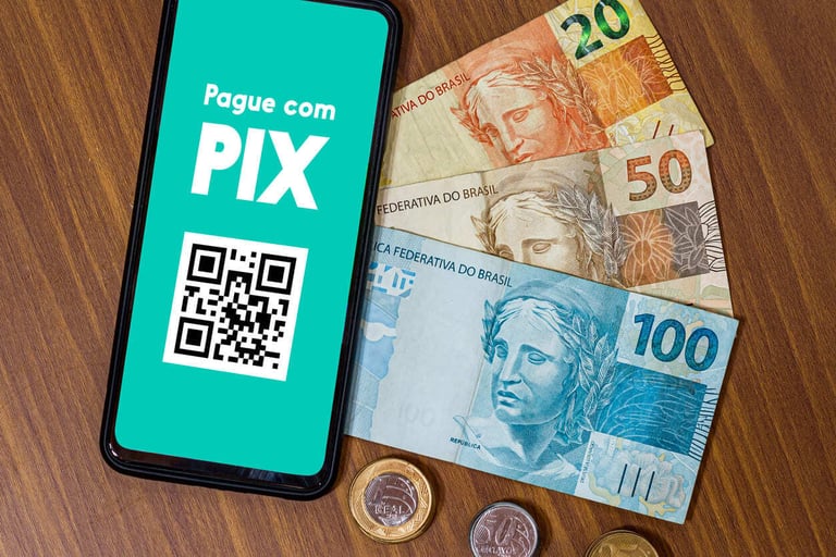 Imagem de um celular na mesa com um QR Code na tela e com a mensagem Pague com Pix. Ao lado, há notas de 20, 50 e 100 reais mais algumas moedas.
