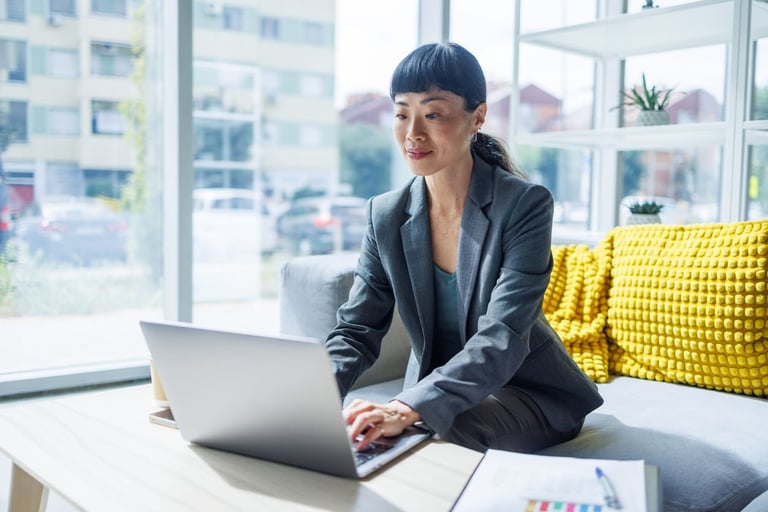 Mulher de negócios usando notebook em escritório moderno com iluminação natural, vestida formalmente e sentada em sofá com almofadas amarelas.