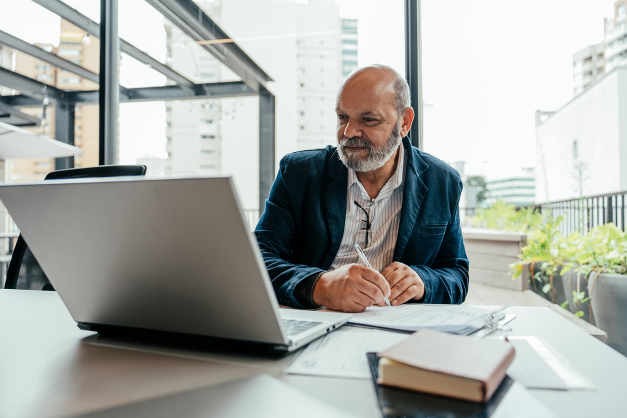 Homem mais velho sentado na frente de uma mesa com um notebook na sua frente e cadernos ao lado. Ambiente de escritório durante o dia.