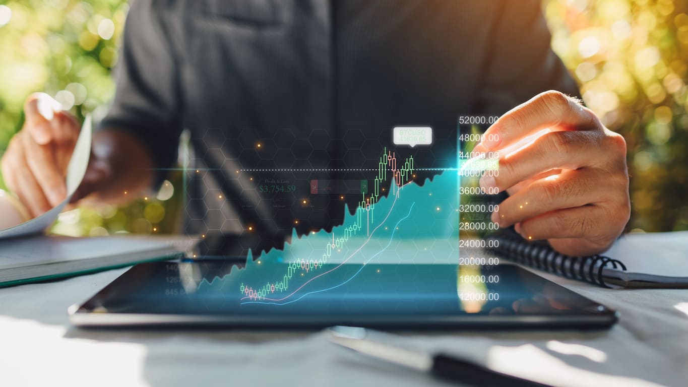 Imagem de um homem mostrando do pescoço para baixo, sentado de frente para uma mesa utilizando um tablet para fazer uma análise financeira. 