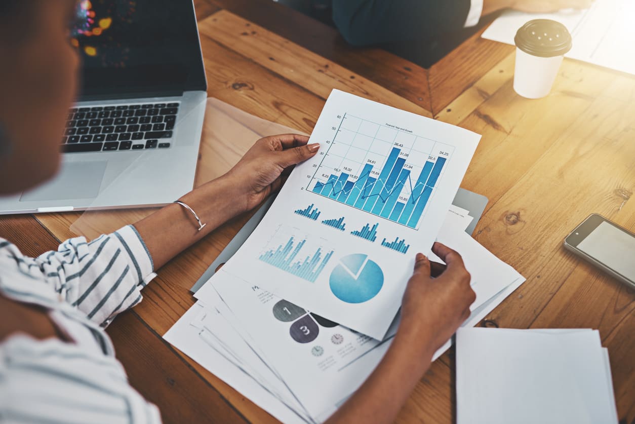 Uma pessoa segurando papéis com gráficos financeiros sobre uma mesa e um notebook aberto ao lado.
